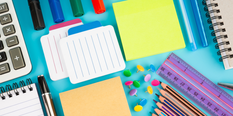 Bird's eye view of stationary set, including thumb tacks, markers, calculator, colored pencils, sticky notes, a ruler, and notebook.