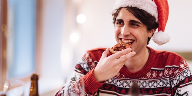 Man wearing a Christmas sweater and Santa hat and eating pizza