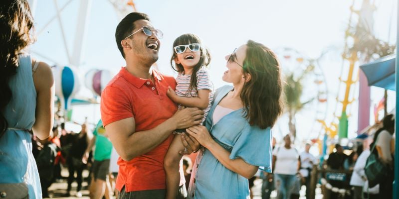 Happy young man and woman holding daughter smiling with sunglasses at a carnival