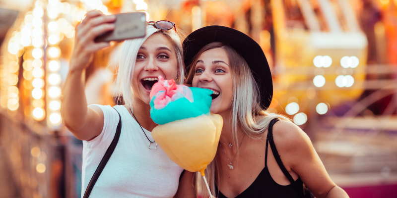 Two women smiling while taking a selfie eating cotton candy while at an amusement park