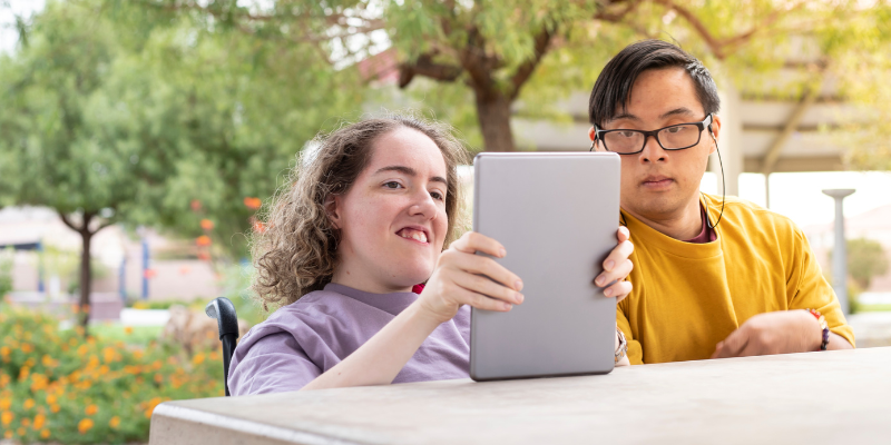 Two people sitting together at a park, looking at a tablet.