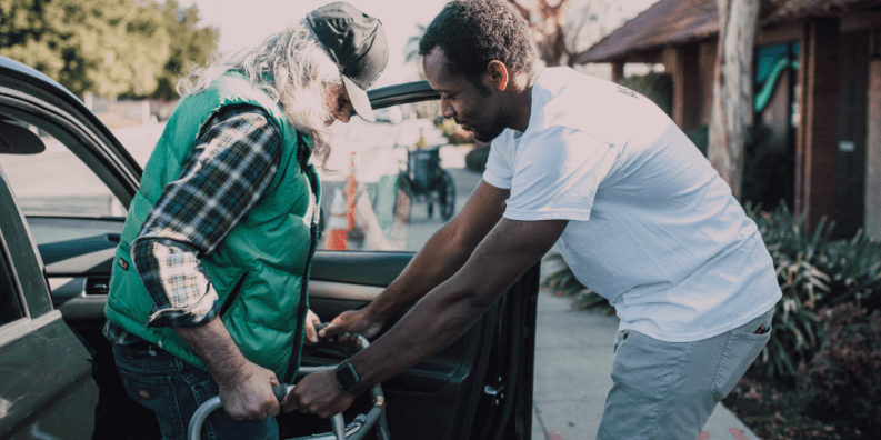 A man helping another man out of a car with a walker.