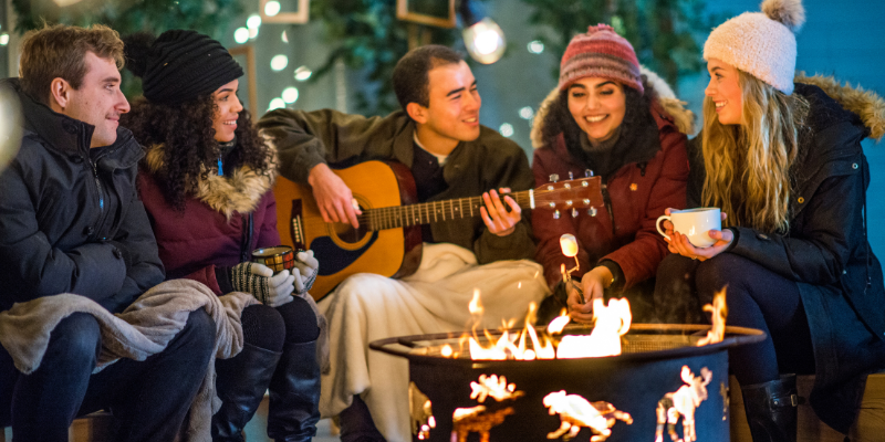 Five people sitting around a campfire. One is playing a guitar.
