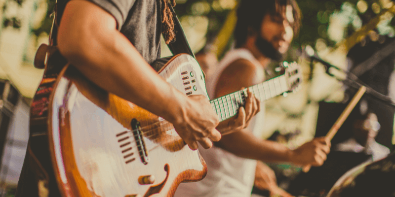 Close up of a pair of hands playing an electric guitar in the foreground with a drummer in the background.  (1)