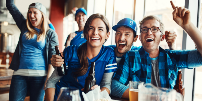 5 sports fans sitting on a couch or standing while cheering.