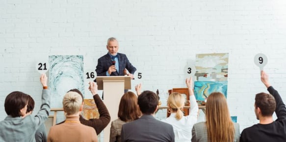 auctioneer taking bids at an art auction while people raise their paddles in the audience