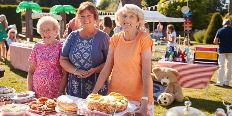 three women smiling at an outdoor bake sale table with desserts displayed in front of them