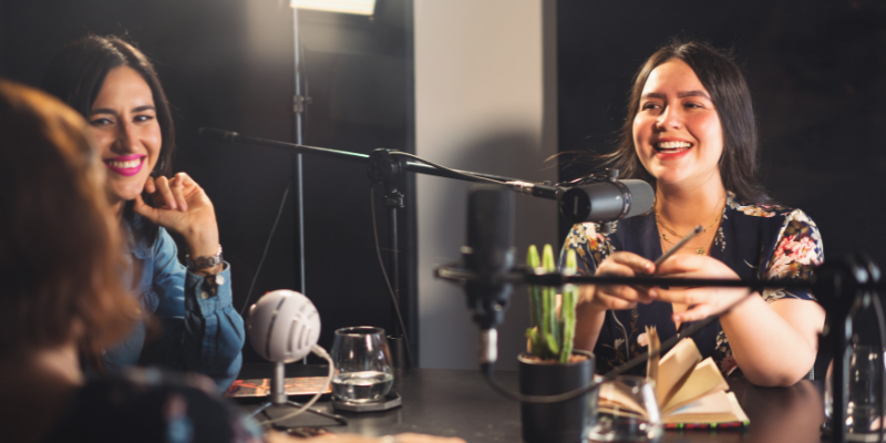 Three women sit at a table with microphones, recording a podcast.