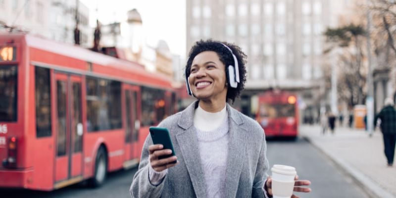 happy woman listening to music on headphones while holding a coffee and phone, city in the background