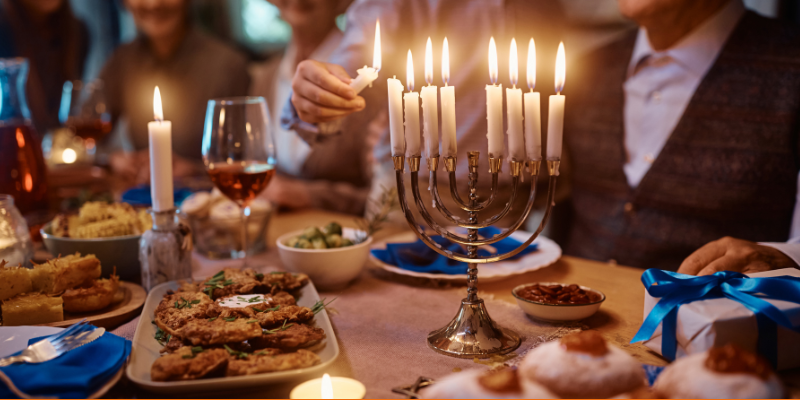 Close-up of Jewish boy lighting the menorah while celebrating Hanukkah with family at the dinner table filled with traditional foods