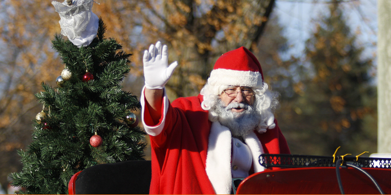 Santa Claus riding in his sleigh waving at Christmas celebrators with a decorated tree in the back of the sleigh