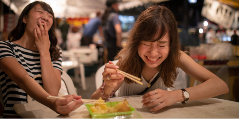 Two young women laughing while eating food with chopsticks at a night festival