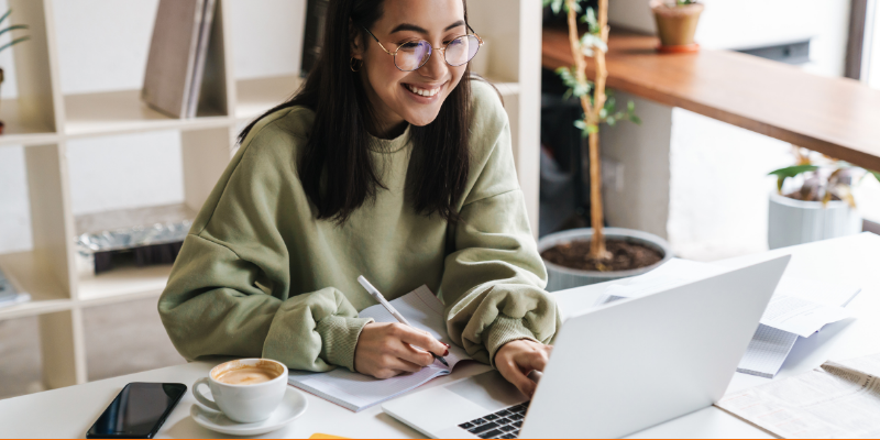 Positive optimistic young student girl with glasses using laptop computer to study indoors