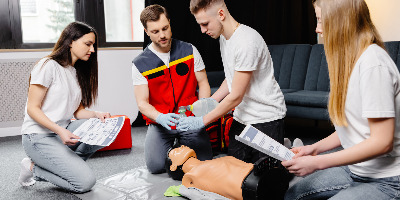 Young man instructor helping to make first aid heart compressions with dummy during group training indoors