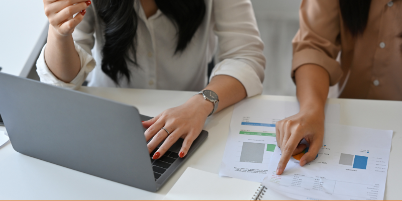 Two business people working in an office reviewing reports on paper and working on laptop