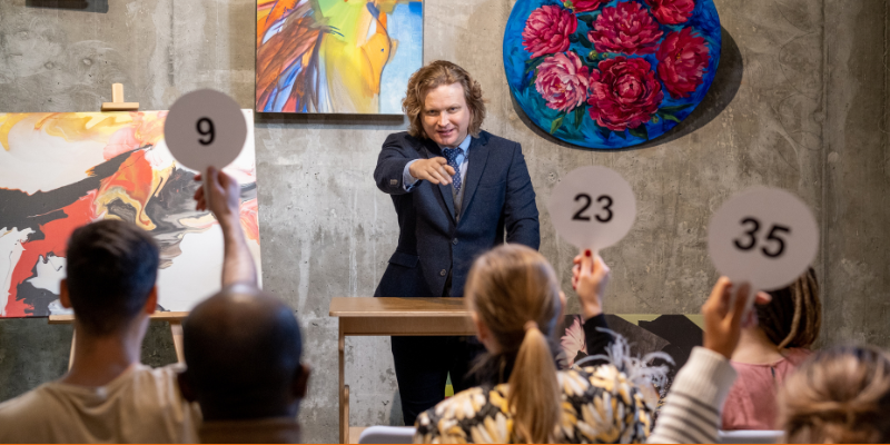 Make auctioneer in a suit standing in front of paintings and pointing at people in the crowd bidding on artwork
