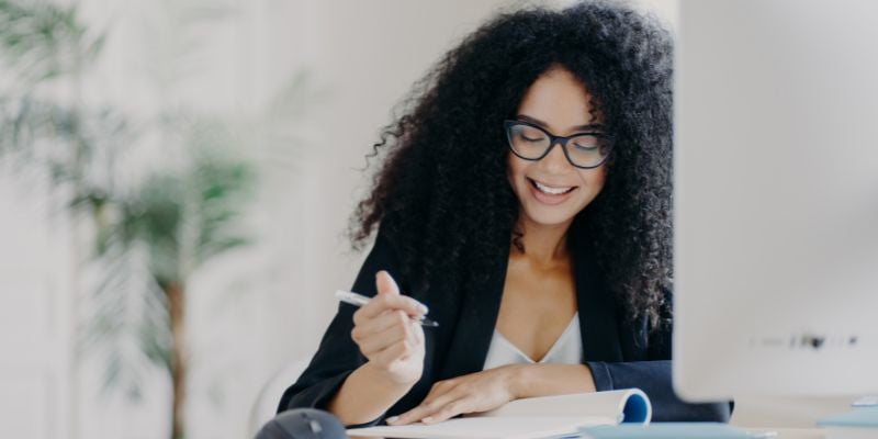 Pleased curly haired woman sitting at desk in front of computer while writing down some information with a pen and paper