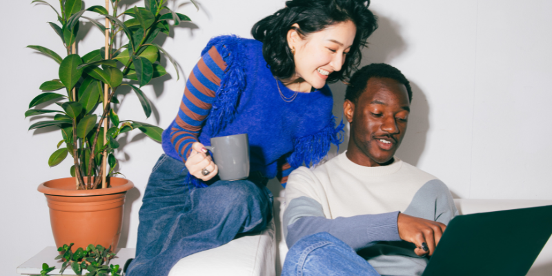 Stylish young woman holding a coffee mug sits on the edge of a chair looking over a casual colleague pointing to something on his laptop