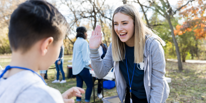 Cheerful young woman gives an elementary-aged child a high five while volunteering at an outdoor event