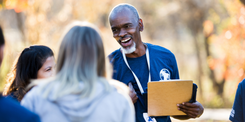 Volunteer wearing a lanyard and holding a clipboard welcoming guests to an outdoor event