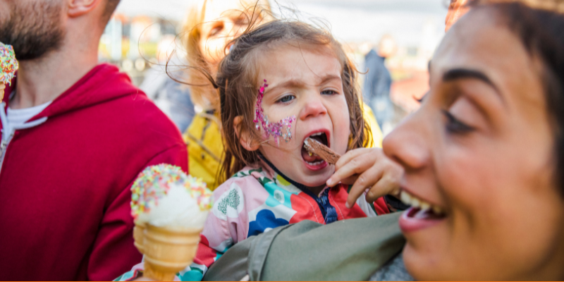 Close up of a young girl with glitter face paint eating an ice cream cone while getting a piggyback ride from her mother