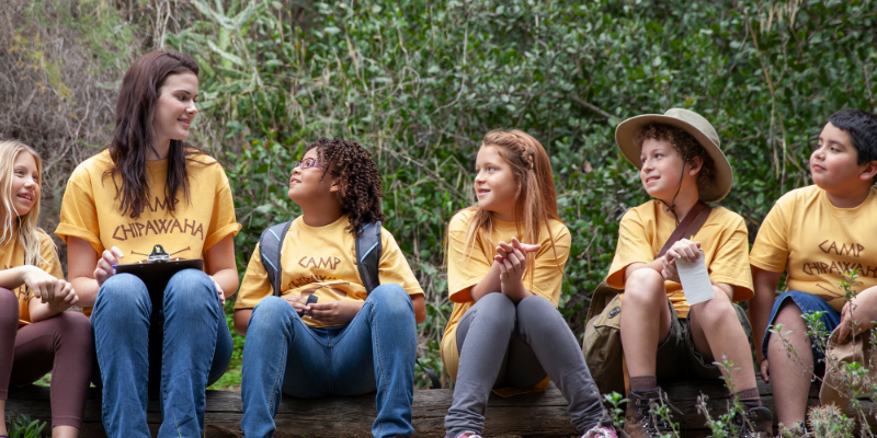 Kids sitting on a log with their camp counselor at a summer camp