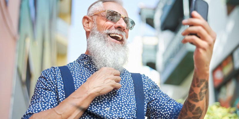 Hip, bearded senior wearing sunglasses and smiling at mobile phone outdoors