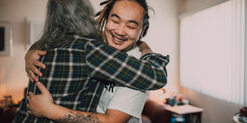 Person in flannel shirt hugging another person in a white t-shirt