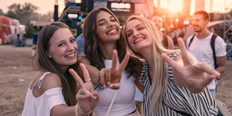 Three female friends smiling and making peace sign at the camera while attending a music festival at sunset