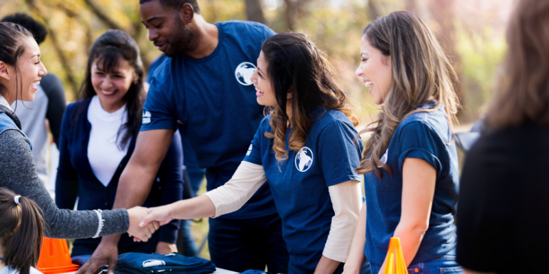 Women shaking hands across a table at a volunteer check-in area for an outdoor event