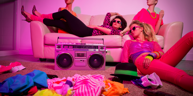 Two girls and a mannequin lounging on a couch wearing 1980s style neon clothing and sunglasses