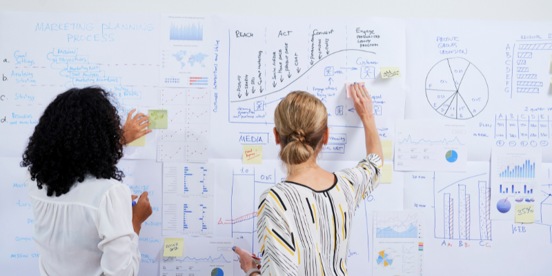 Rear view of two female business executives reviewing charts with paper and post-its stuck onto the wall