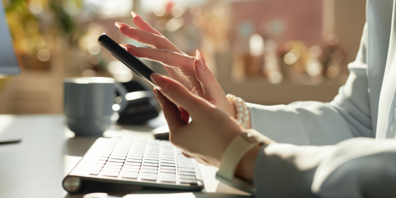 Side view of female hands scrolling on phone with a keyboard and coffee mug on the desk in background
