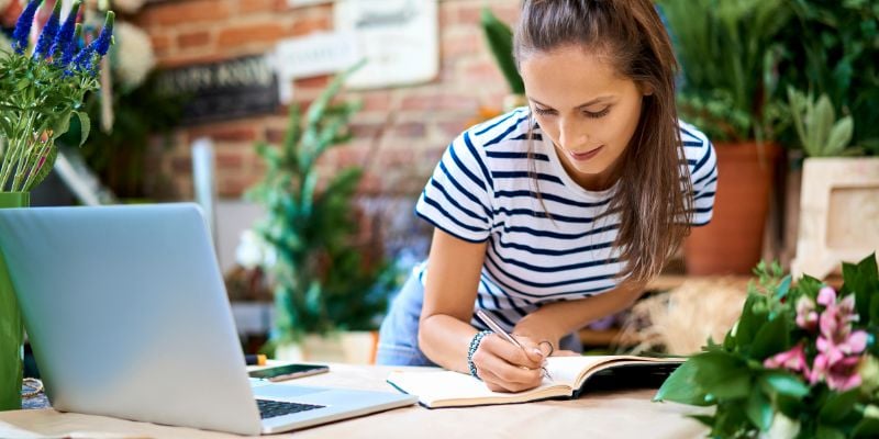 woman writing in a notebook next to an open laptop, standing in a flower shop 