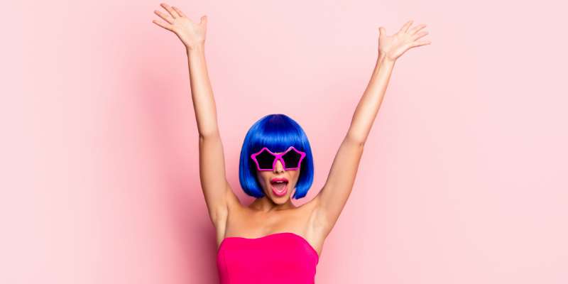 woman with blue wig and star sunglasses wearing bright pink dress in front of a light pink background