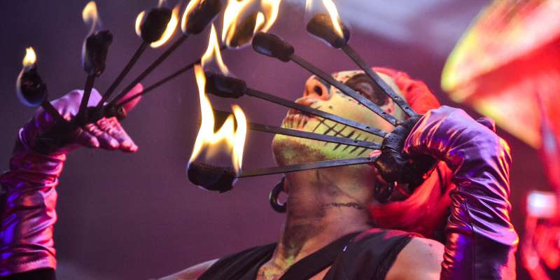 circus performer with skull makeup blowing on multiple fire batons held in front of their face