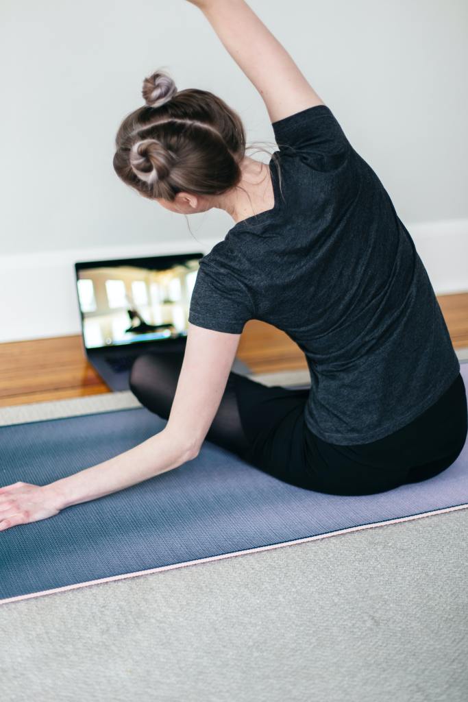 woman with hair in two buns side-bending while sitting on a yoga mat in front of laptop