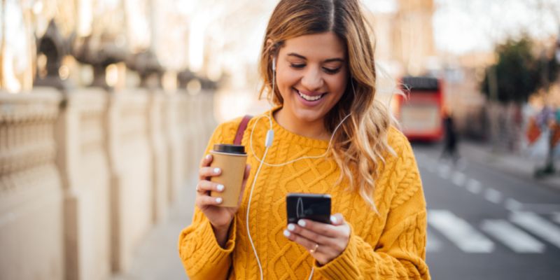 smiling woman outdoors wearing a yellow sweater holding a to-go coffee in one hand and a mobile phone in the other