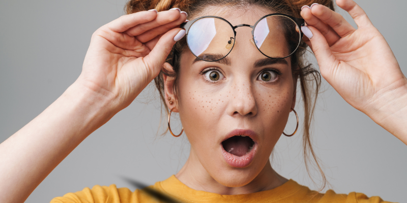 Close-up shot of a cute young woman looking surprised the lifting glasses off her eyes on a gray background