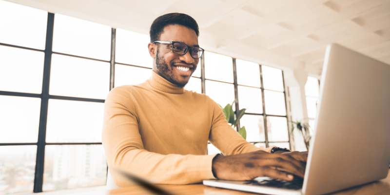 Smiling African American guy wearing glasses using a laptop in a modern office with lots of windows