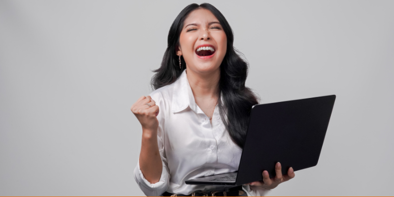 Happy young Asian woman raising her first in excitement while holding laptop computer against a gray background