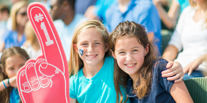 Two young girls smiling into camera holding a foam finger at a sporting event