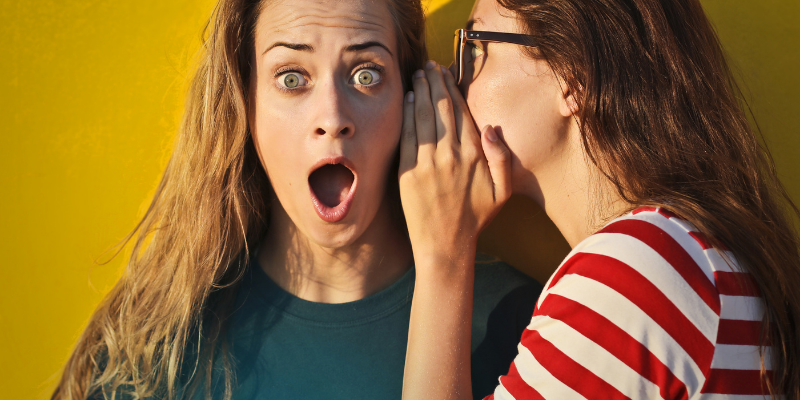 Girl whispering into her friends ear against a yellow background