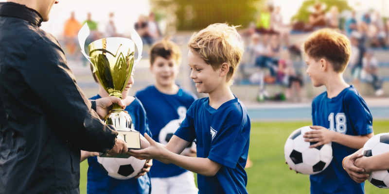 Coach handing soccer trophy to a young boy surrounded by teammates on a soccer field