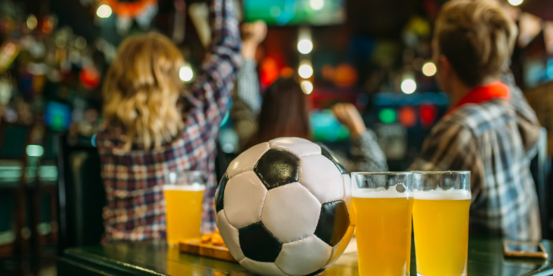 Soccer ball and beer on a table in a sports bar with fans cheering in the background