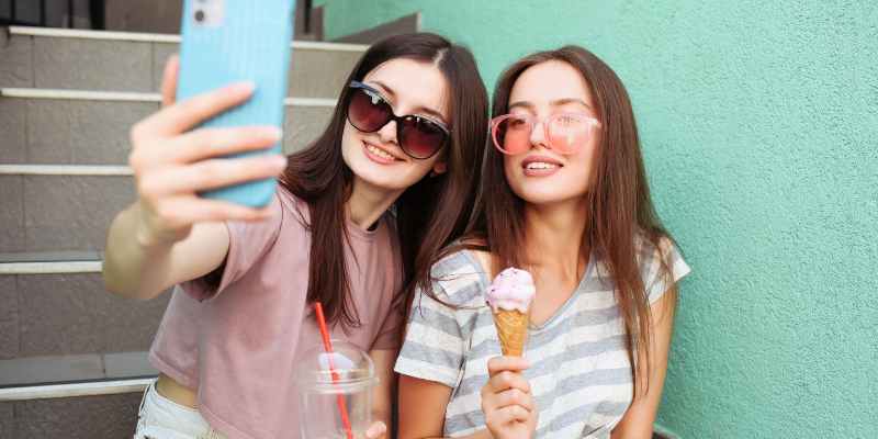 Two women sitting down taking a photo with an ice cream and drink in their hand