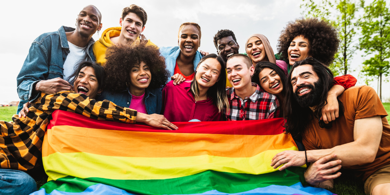 Group of twelve people of varying races and ethnicities standing or sitting in front of a rainbow pride flag.