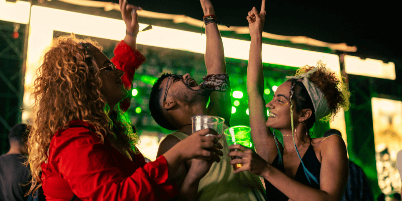 Three people raising their arms and drinks in the air as they dance at a concert.